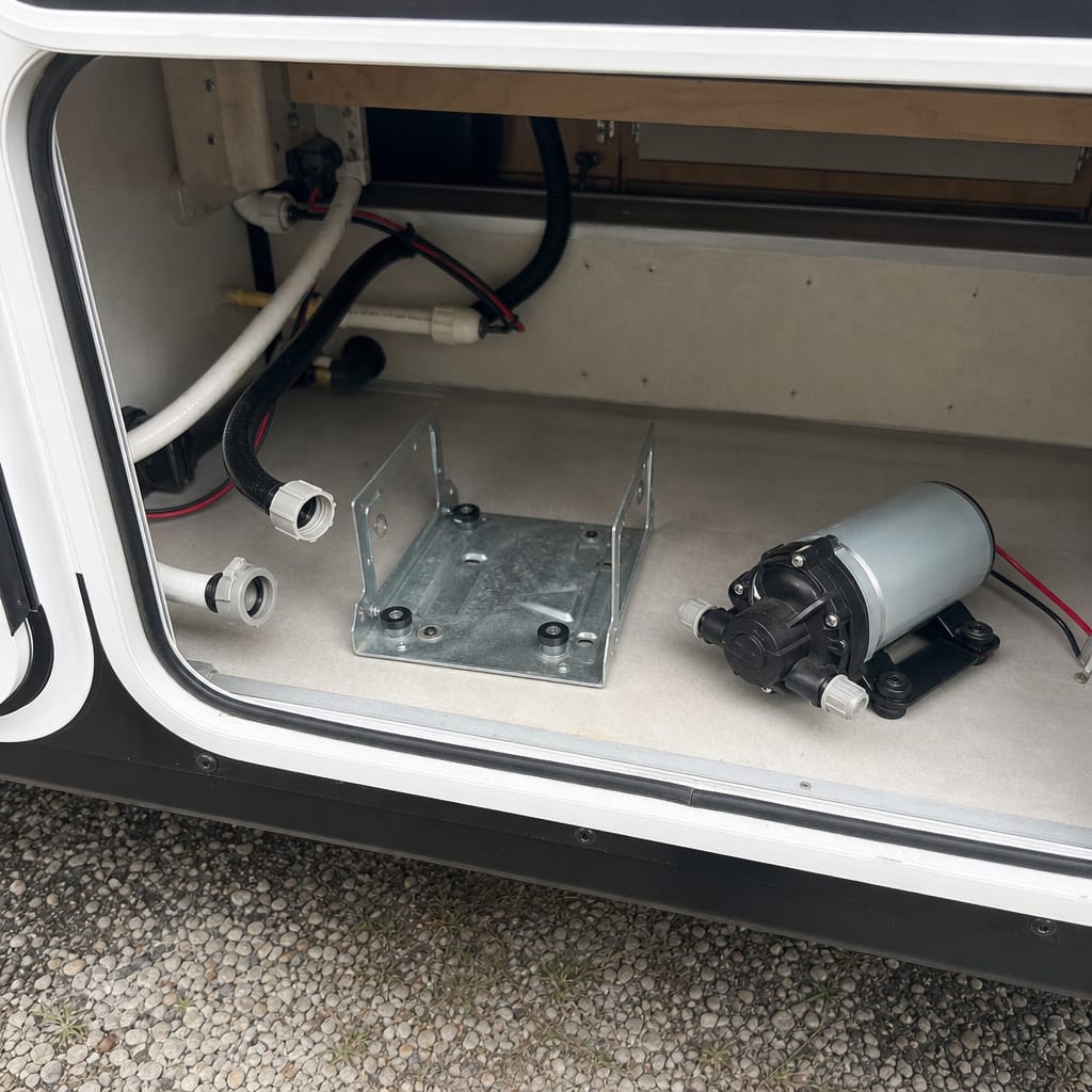 RV water pump compartment with the old pump removed and a new unbranded replacement pump set beside the mounting bracket, daylight" (139 chars — see note below)